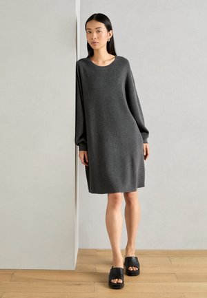 Gray oversized sweater dress with long sleeves, round neckline, and ribbed cuffs, paired with black flat slide sandals. Wooden flooring background.