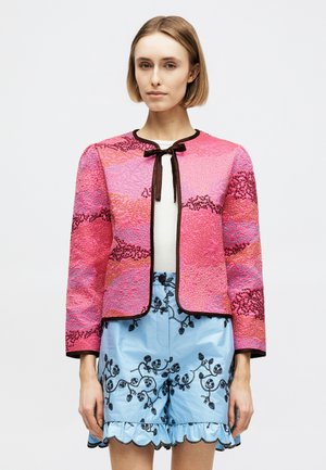 Femme portant une veste rose texturée avec bordure noire et nœud sur un haut blanc, associée à un short bleu clair brodé aux bords festonnés.