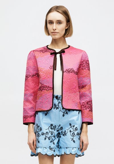 Femme portant une veste rose texturée avec bordure noire et nœud sur un haut blanc, associée à un short bleu clair brodé aux bords festonnés.