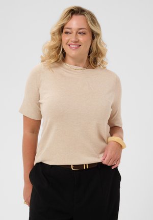 Femme souriante aux cheveux blonds bouclés portant un haut beige à manches courtes, un pantalon noir, un collier en or, un bracelet beige et une ceinture marron.
