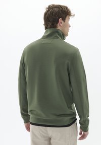 Pullover lungo verde oliva con maniche lunghe e colletto alto, caratterizzato da un tessuto testurizzato e polsini a costine, dettagli di cucitura visibili e una vestibilità comoda.