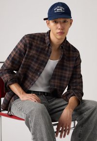 Καρό πουκάμισο σε καφέ και ναυτικό μπλε με κούμπωμα κουμπιών, σε συνδυασμό με λευκό φανελάκι και γκρι τζιν παντελόνι. Ναυτικό μπλε καπέλο με λευκή λεπτομέρεια κειμένου.