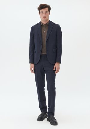 Marineblauw pak met een slim fit, gecombineerd met een bruine polo. Het pak heeft een gestructureerde stof en beschikt over notch revers en subtiele zakken. Zwarte loafers.