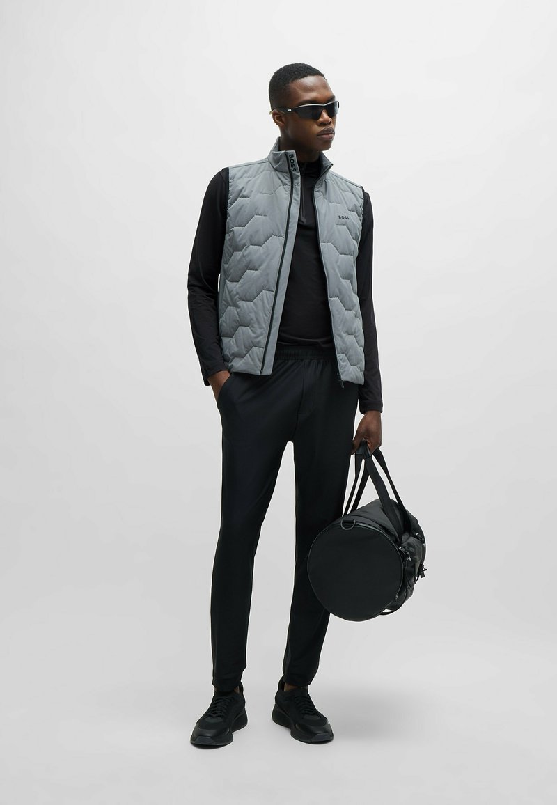 Chaleco acolchado gris sobre una camiseta negra de manga larga, combinado con pantalones negros ajustados y zapatillas. Sosteniendo un bolso de viaje redondo negro.