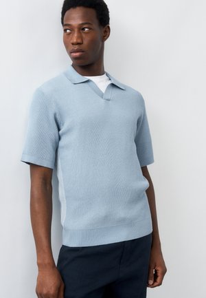 Άνδρας που φοράει γαλάζιο πλεκτό πόλο με κοντά μανίκια και σκούρο παντελόνι, κοιτάζει προς τα αριστερά του μπροστά σε απλό ανοιχτό φόντο.