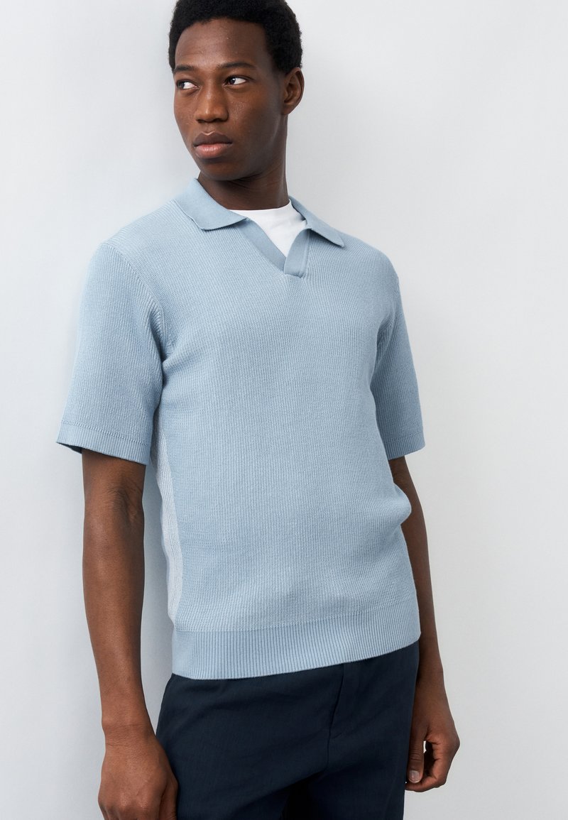 Άνδρας που φοράει γαλάζιο πλεκτό πόλο με κοντά μανίκια και σκούρο παντελόνι, κοιτάζει προς τα αριστερά του μπροστά σε απλό ανοιχτό φόντο.