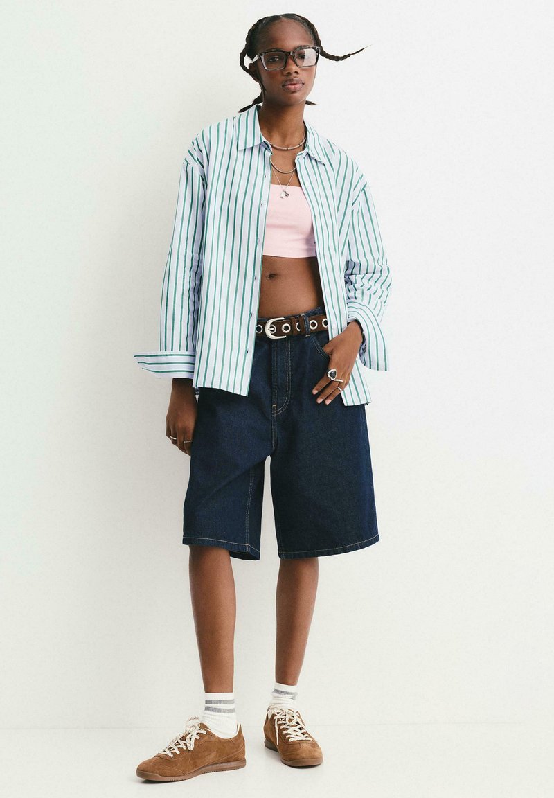 Giovane donna con capelli intrecciati che indossa occhiali, camicia a righe, top corto rosa, pantaloni corti in denim scuro, calze bianche e sneakers marroni, in piedi contro un muro bianco.