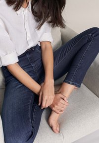 Jeans de mezclilla oscuros de tiro alto con una textura suave, combinados con una camisa blanca de cuello, usados mientras se está sentado en un sofá de color claro.