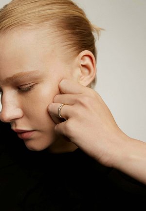 Anillo de oro llevado en la yema de un dedo, con un diseño elegante y minimalista. La mano descansa contra la cara con piel suave y un fondo neutro.
