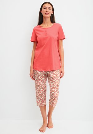 Vrouw staat blootsvoets, draagt een koraalkleurige korte mouw top met knoopdetails en een korte broek met bloemenpatroon tegen een witte achtergrond.