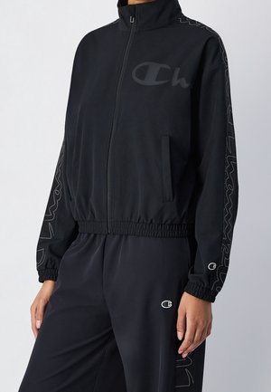 Persona con chaqueta negra con cremallera y pantalones a juego con puños elásticos, con logos sutiles de Champion en el pecho, las mangas y los pantalones.