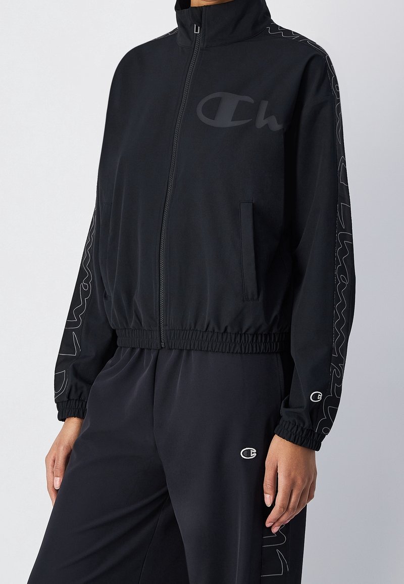 Personne portant une veste noire zippée et un pantalon assorti avec des poignets élastiques, avec des logos Champion discrets sur la poitrine, les manches et le pantalon.
