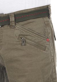 Olivgrüne Shorts aus strukturiertem Stoff, mit einer Reißverschlusstasche, genähten Akzenten und einem kontrastierenden grünen Gürtel mit roter Stickerei.