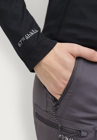 Schwarzlangärmliges Shirt mit dem Logo "67°N'ähkä" auf dem Ärmel; graue Hose mit Reißverschlusstasche und Kordeldetail am Bund.