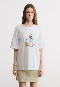 Νεαρή γυναίκα που φοράει λευκό, φαρδύ T-shirt με κινούμενο σχέδιο μιας ξανθιάς κοπέλας που βγάζει βόλτα έναν κίτρινο σκύλο, συνδυασμένο με μπεζ φούστα.