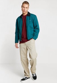 Tropfenauswahl Jacke mit schwarzem Kragen, darüber ein kastanienbraunes Hemd, kombiniert mit beigen Hosen und schwarzen Sneakers. Schlichtes Design, lässiger Schnitt.