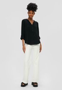 Chemise noire à manches longues avec un col en V et des détails de boutons, associée à un pantalon blanc évasé et des chaussures à talons épais noires. Tissu texturé.