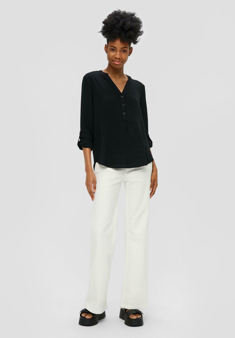 Chemise noire à manches longues avec un col en V et des détails de boutons, associée à un pantalon blanc évasé et des chaussures à talons épais noires. Tissu texturé.