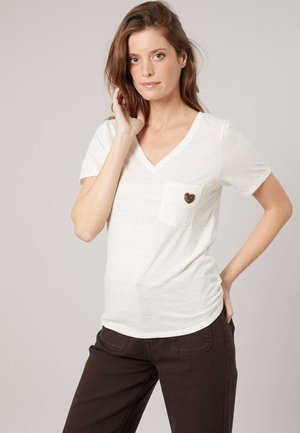 Femme portant un t-shirt blanc à col en V avec un petit cœur sur la poche poitrine, et un pantalon foncé, posant avec une main sur la hanche.