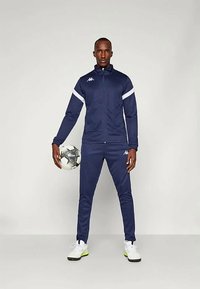 Navy treningsdrakt med hvite detaljer, som inkluderer en jakke med glidelås og slim-fit bukser. Tilbehør inkluderer en svart-hvit fotball og hvite sko.