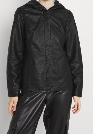 Femme portant une veste à capuche noire en simili cuir à manches longues et un pantalon large en simili cuir noir.