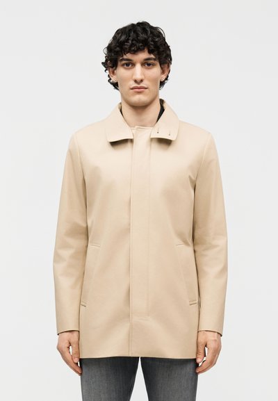 Jeune homme aux cheveux bouclés noirs portant un manteau long beige et un jean foncé, debout droit devant un fond gris clair uni.