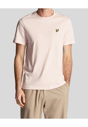Mężczyzna noszący jasnoróżowy t-shirt z krótkim rękawem z małym żółtym logo ptaka na piersi i beżowe spodnie, jedna ręka w kieszeni, neutralne tło.