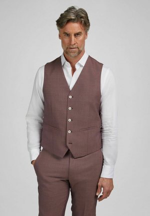 Man met grijs haar, gekleed in een wit overhemd en een op maat gemaakt mauve-colbert met bijpassende broek, staand tegen een effen achtergrond.