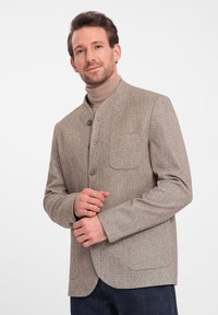 Braune Fischgrätjacke mit Stehkragen, vier Knöpfen und einer Brusttasche. Über einem hellbraunen Rollkragenpullover getragen, kombiniert mit dunklen Jeans.
