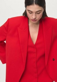 Woman looking down wearing a bright red buttoned vest and oversized red blazer against a plain light background.