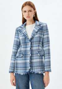 Blå och vit tweedblazer med en strukturerad design, fransade kanter, två framfickor och två knappar framtill. Buren med en vit topp.