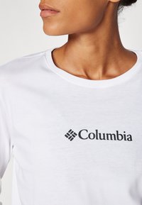 Bijela pamučna majica s okruglim izrezom i crnim logotipom "Columbia" na lijevoj strani prsnog područja. Glatka tekstura s klasičnim krojem.