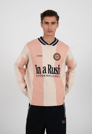 Ung mand iført en lyserød og cremefarvet stribet sweater med teksten "In a Rush International" og en cirkulær logo, kombineret med sorte bukser.