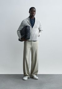 Homme debout contre un mur uni portant un pantalon large beige, un polo bleu marine, une veste zippée grise, des baskets blanches, tenant un sac matelassé noir sous le bras.