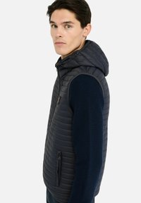 Veste matelassée bleu marine avec capuche, tissu texturé, poche à fermeture éclair à l'avant, et manches en tissu tricoté contrastant.