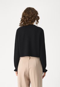 Melna, īsā džemperis ar garām piedurknēm un viļņainām aprocēm, izgatavots no mīksta materiāla. Uzvilkts pār bēšām, augstā jostasvietas biksēm.