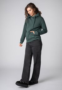 Groene katoenen hoodie met verstelbare koorden, een voorkantzak en logo-detail. Combinatie met zwarte wijde jeans en zwarte instap-schoenen.