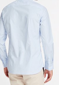 Uomo che indossa una camicia a maniche lunghe azzurro chiaro e pantaloni beige, visto da dietro su uno sfondo bianco semplice.
