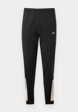 Black sweatpants with green side stripes and beige accents at the cuffs. Elastic waistband and Lacoste logo on the front. Smooth texture.