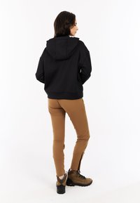 Zwarte hoodie met een losse fit, gecombineerd met tan, tapse broek met ritsen bij de enkels, aangevuld met bruine wandelschoenen.