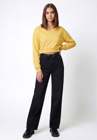 Dzeltena īsa džemperis ar V veida izgriezumu, kombinēts ar augstu jostasvietu melnām džinsa biksēm un melnām ādas kurpēm ar šņorēm, papildināts ar teksturētu jostu.