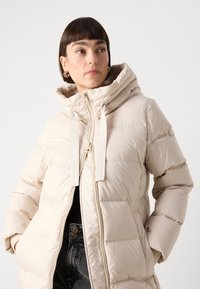 Pufferjack in lichtbeige, met een capuchon, ritssluiting en elastische trekkoorden. Textuur met geborduurde delen.