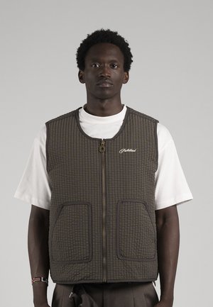Un homme porte un gilet sombre à carreaux, zippé sur le devant et doté de deux grandes poches, par-dessus un T-shirt blanc, sur un fond gris uni.