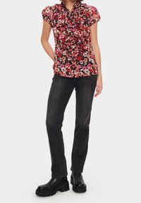 Blouse à fleurs noire, rose et rouge avec un col à volants et des manches courtes, associée à un jean foncé et des bottes noires épaisses.