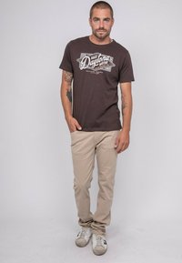 T-shirt en coton marron avec un imprimé graphique blanc "Daytona", associée à un pantalon chino beige et des baskets blanches avec des accents gris.