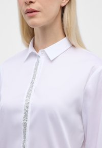 White blouse featuring a classic collar, front button placket, and a decorative silver sequined stripe down the center. Smooth fabric texture.
