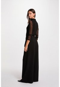 Vrouw in zwarte wijde broek en een zwart top met ondoorzichtige, patroon sleeves, staand in een driekwart pose tegen een effen achtergrond.
