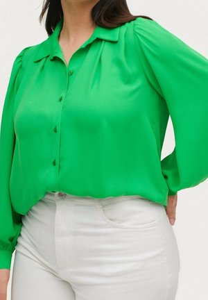 Femme portant un chemisier boutonné à manches longues vert vif, rentré dans un pantalon blanc taille haute, main posée sur la hanche.