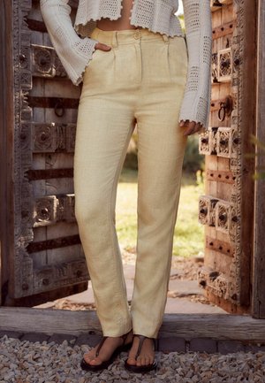Pantalons en lin jaune clair avec une coupe décontractée et des plis à l'avant. Associés à un cardigan en tricot blanc texturé et cropped, ainsi qu'à des sandales simples.