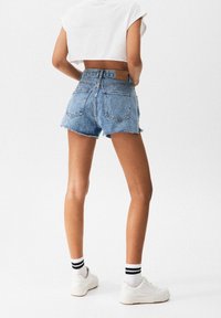 Mujer con pantalones cortos de mezclilla azul desgastados, top corto blanco, zapatillas blancas y calcetines blancos con rayas negras, de pie contra un fondo liso.